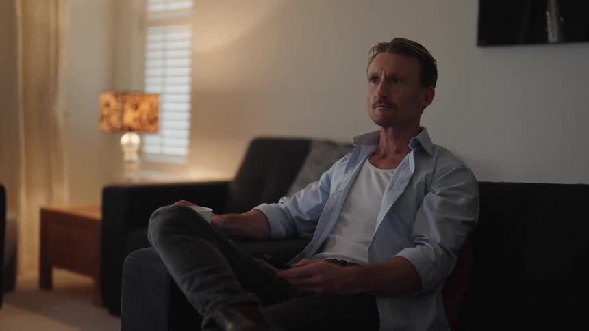 Man sits in living room drinking from a mug and watching televison