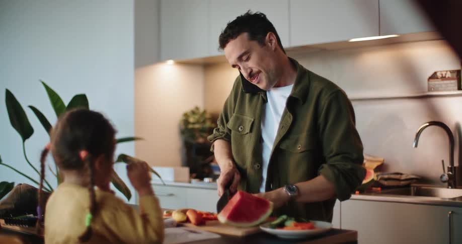 Daughter with braids and yellow sweater watches father with mustache green and white shirts cutting vegetables at kitchen island while talking on mobile phone