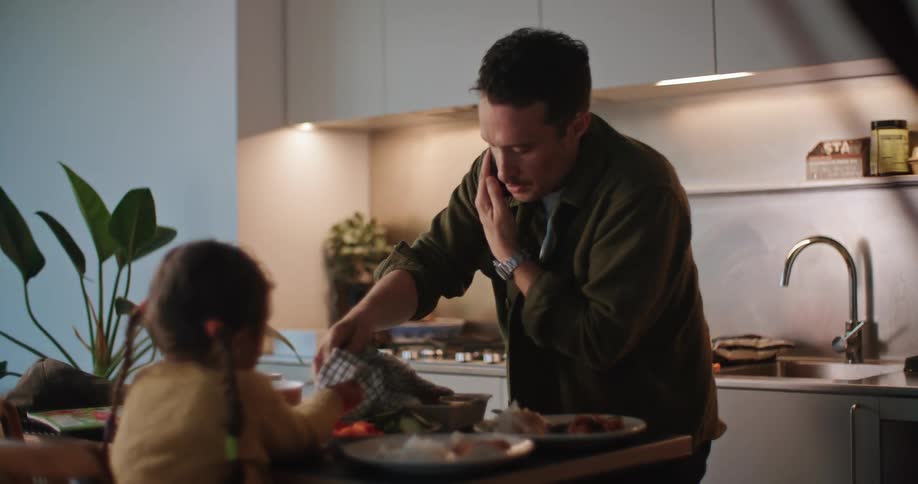 A man talks on the phone while trying to cook for his young daughter