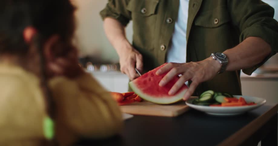Daughter with braids and yellow shirt watches father with mustache green shirt and white top cuts fruits and vegetables while talking on smartphone in the kitchen