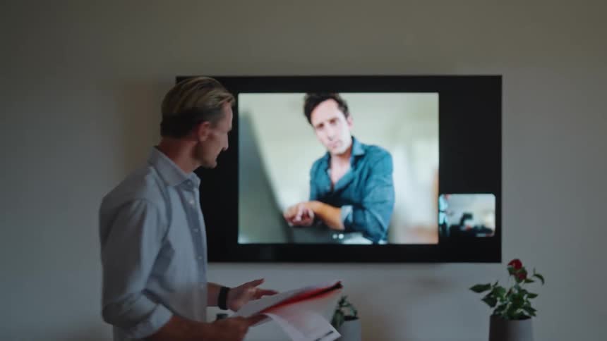 A man holding papers as he talks to a person on video chat on the tv screen