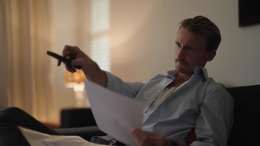 A man changes tv channels with a remote while reading papers on the couch