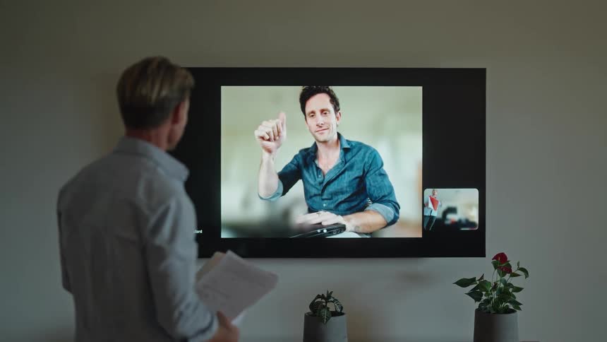 A man is standing in a room waving to a man on the big screen on his wall as they finished having a video conference