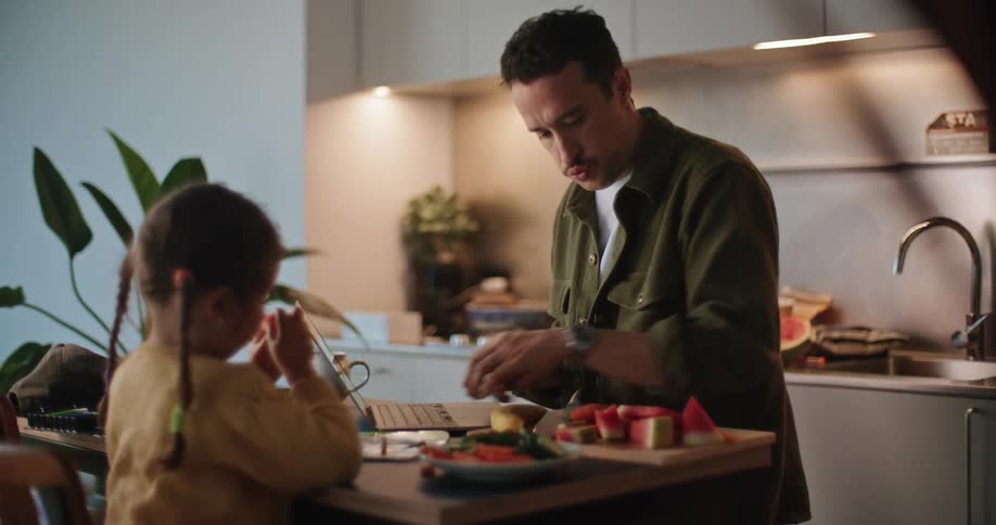 A father is standing at a tall kitchen table working on a laptop while at the same time talking to his young daughter and handing her cut watermelon pieces as she is sitting in a chair across from him