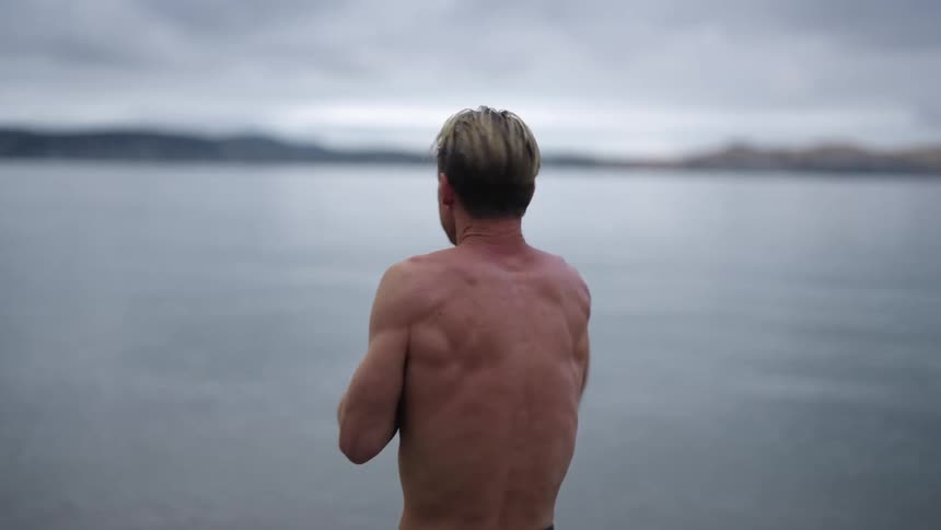 A bare chested man is standing along the shoreline looking at the water and rubbing his hands together as he appears to be cold