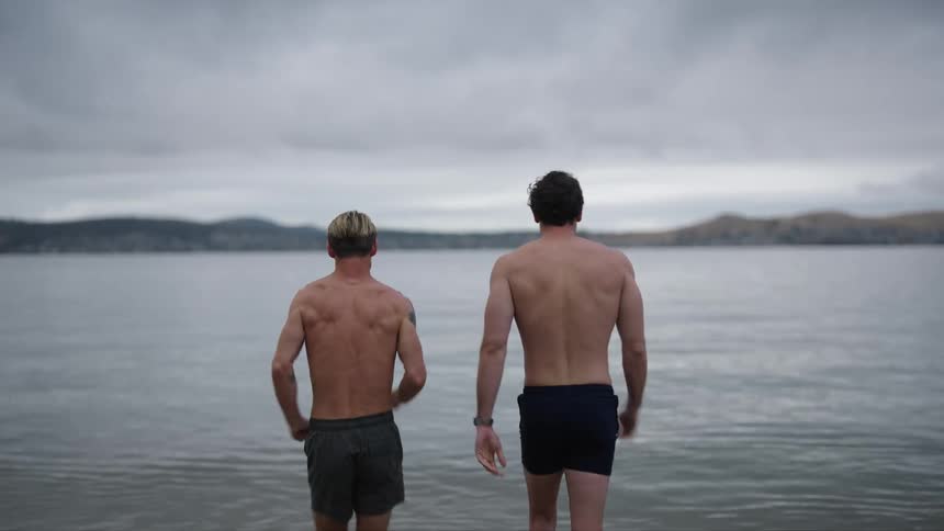 A pair of men in swim trunks bump fists before walking into a lake
