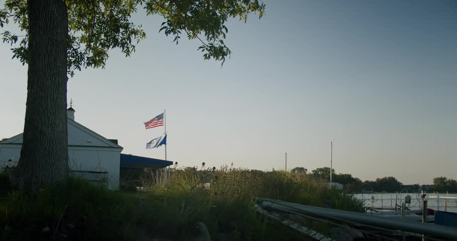 Flags wave in breeze over building surrounded with overgrown lawn near lake
