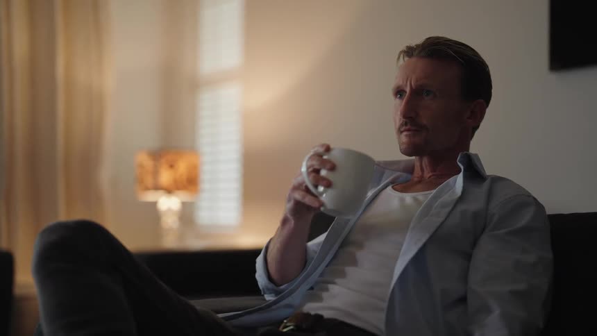 Man sits down on the couch at home to watch television while drinking a hot beverage
