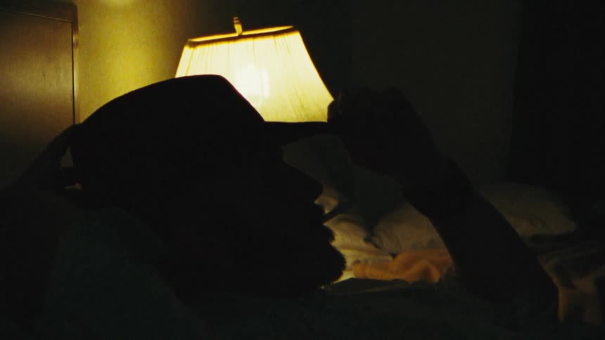 Man with hat lying on bed and smoking a cigarette is being silhouetted against the bedside lamp