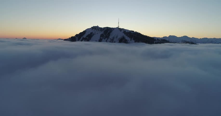 Aerial view of an antenna atop a mountain top surrounded by clouds at evening