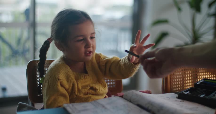 A little girl takes a crayon from someone and draws on the book in front of her