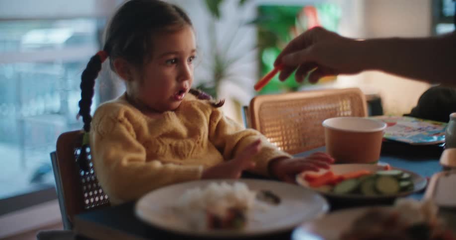 Little girl sitting at the dinning table eats carrots sticks with the help of her dad