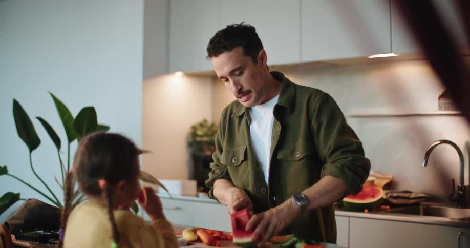 Father slicing watermelon as young daughter eats it at kitchen counter