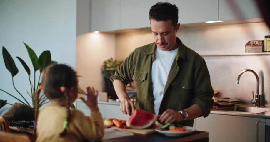 A father slices watermelon for his daughter sitting across from him in the kitchen