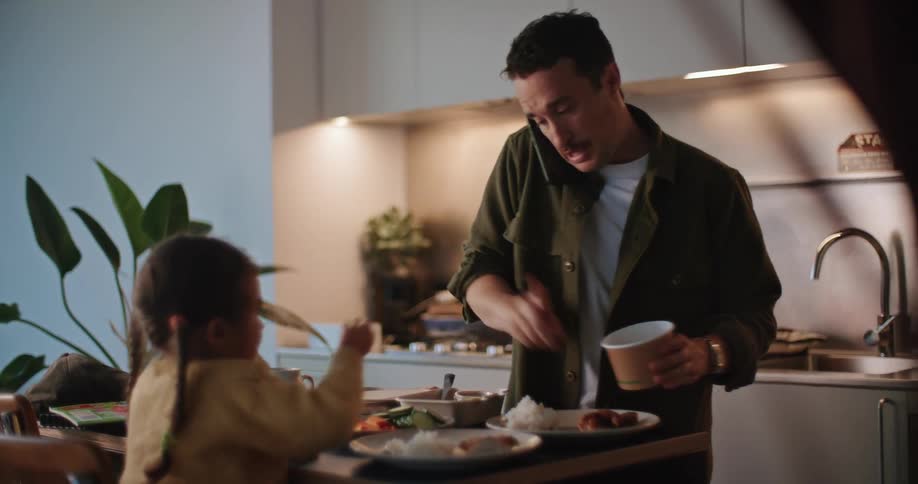 A father prepares food for his daughter sitting in front of him as he talks on the phone
