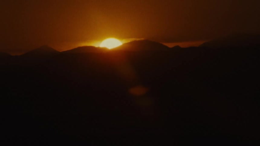 A bright sun sets over mountains in a dark red sky