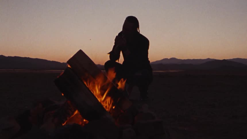 A silhouette of a man sitting next to a fire at dusk with mountains in the background