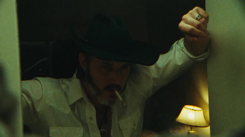 A man puts on a hat while smoking a cigarette walks up to a mirror and throws the cigarette at it
