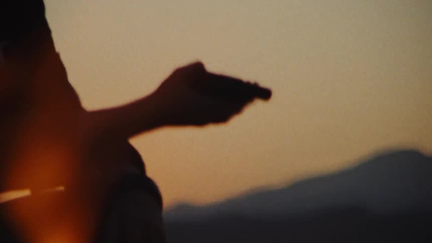 Silhouette of a man handling a revolver while smoking sitting near a campfire at evening