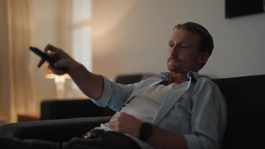A man sitting on his couch using a remote yawning and then crossing his arms