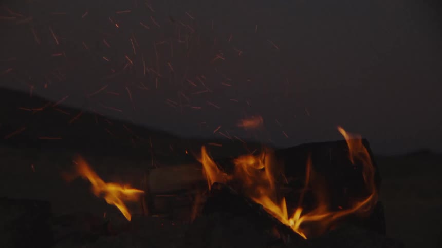A man kicks the logs of a campfire as sparks rise among the fire and flames