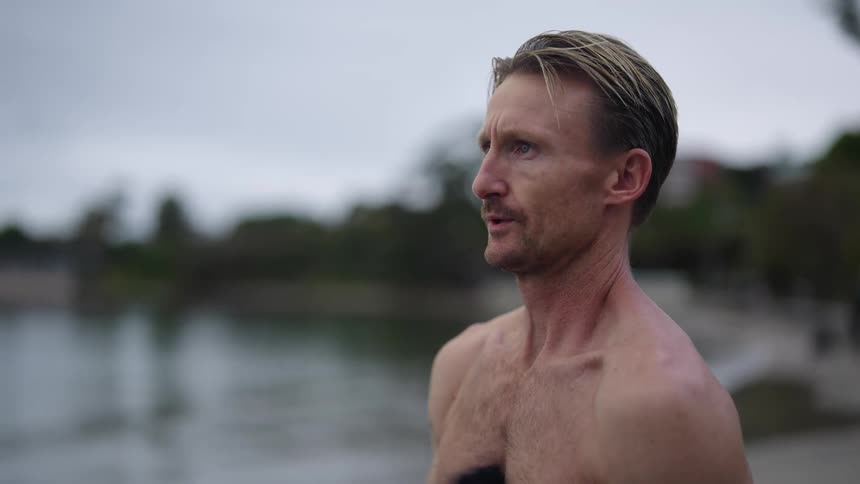 A man is standing along the shoreline as he takes off his jacket and stands bare chested looking at the water