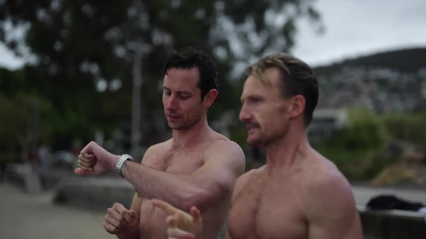 Two bare chested men are standing outside as one man looks at his wristwatch they appear to be cold and he hugs the other man and they laugh