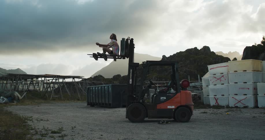 Young man sitting on the raised forks of a forklift parked on a yard at morning