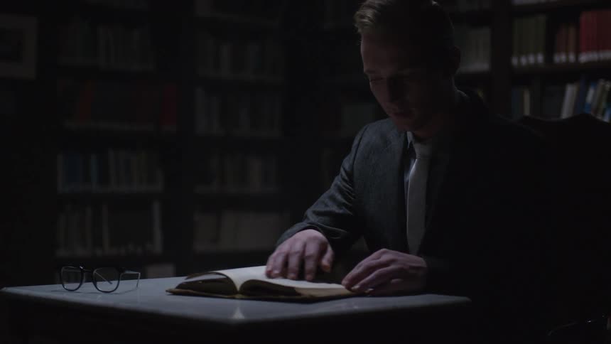 Man opening a book in a library and turning the pages while sitting at a small table
