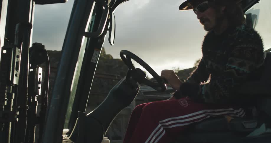 Man with long hair drives a forklift through a yard at morning