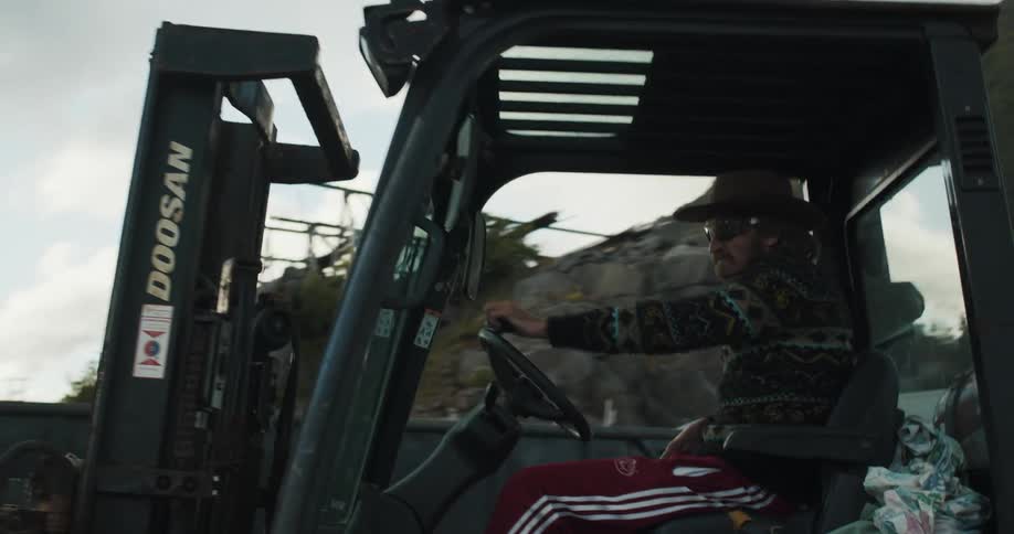 A man in a cowboy hat and sunglasses is laid back with one hand on the wheel driving a forklift with a pallet on it from one area of a construction site to a spot not far away