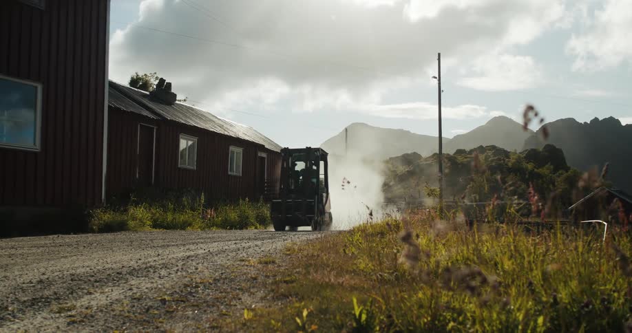 Man drives forklift along dirt road between building and field with mountains in the background