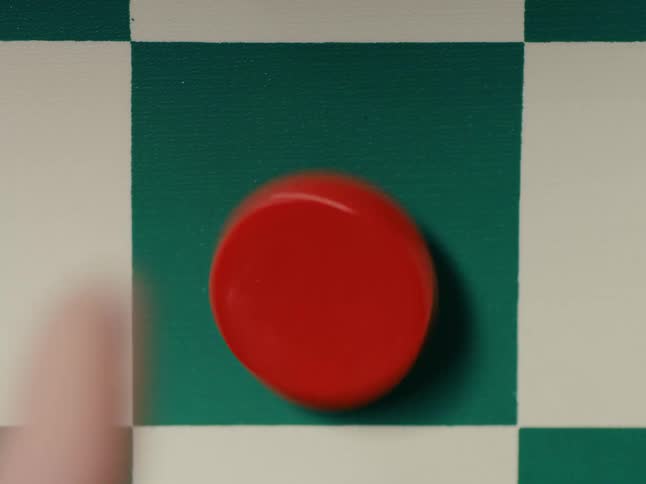 A red checker piece sits on a green square on the checker board as a person stands it on the edge and flips it over