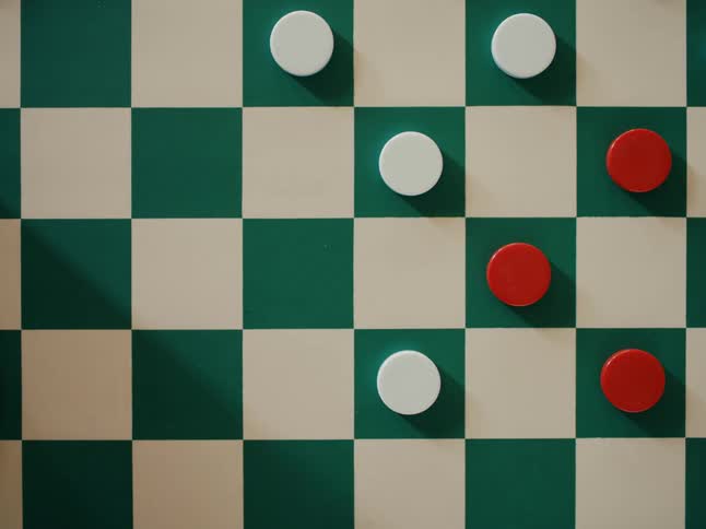 A player in a checker game uses their finger to push a red game piece to a green square and a hand is moving pieces and taking them off the game board