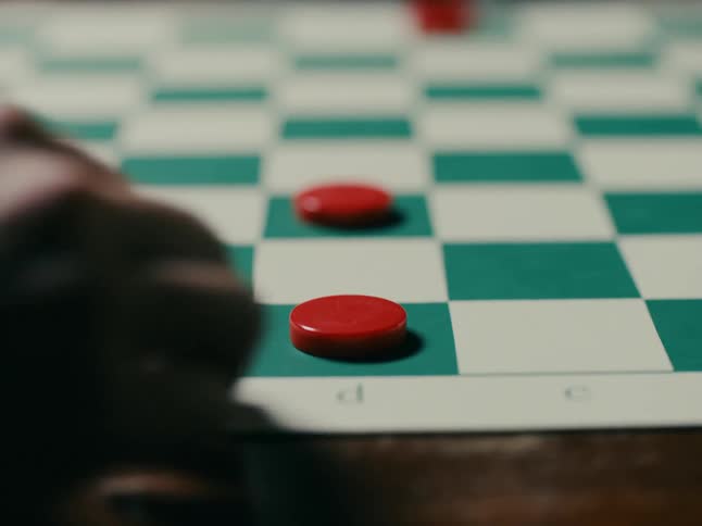 A person moves a red chip on a checkered board while another person puts a red chip on top of the first
