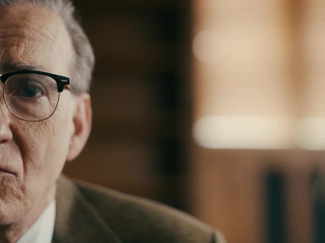 A man is wearing a suit and glasses while sitting indoors