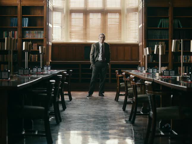 Professor stands between rows of desks on a university library looking at the camera
