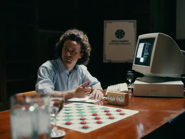 Man sitting on a desk in front of a computer and a checkers board waits patiently