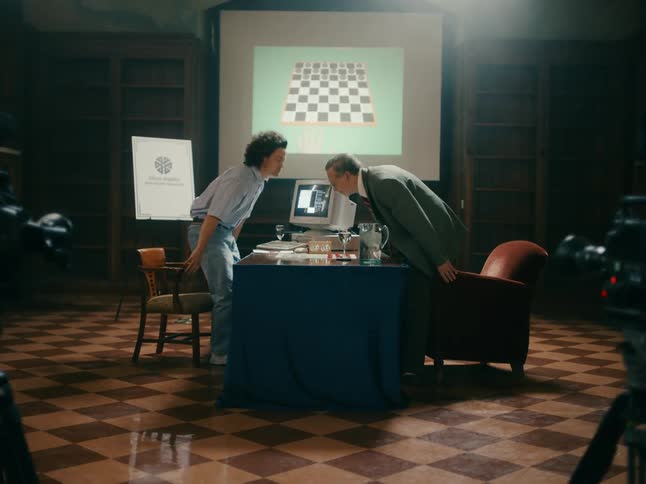 Two cameras record computer researcher interacting with older man in regards to a checker game and using an old computer