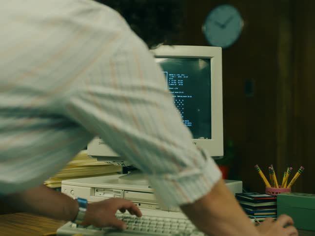 A man is standing over an old computer with a checker game on the monitor as he types on the keyboard