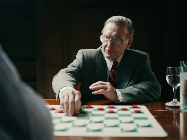 A man steps up to a table with a checkerboard set up as he pulls out his chair he reaches his hand out to shake hands with his opponent and sits down as the game begins