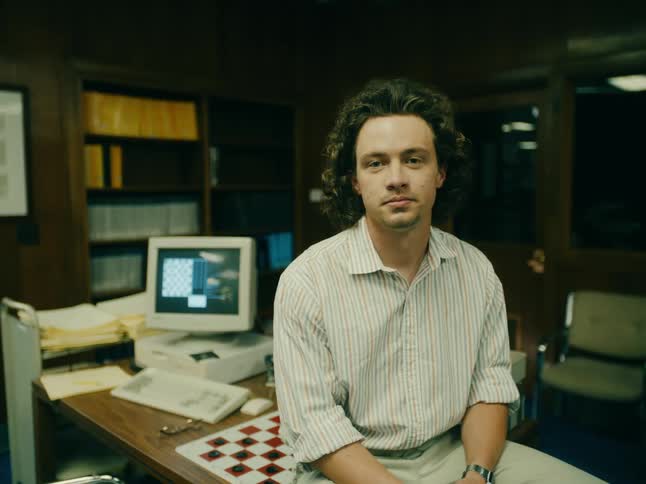 A man sits on his desk with a computer and game board on the desk behind him