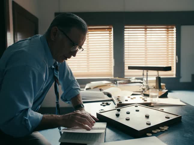 School teacher sitting at desk in classroom alternately playing checkers and reading book