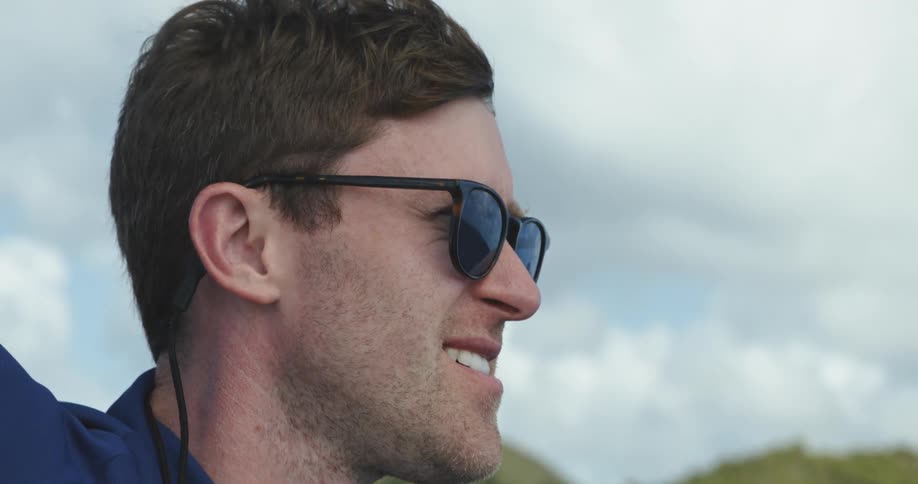 A man is standing with one arm up holding onto something beside him as he is slowly rocked up than down all the while the wind blows his hair back and he squints under his sunglasses under a cloudy blue sky