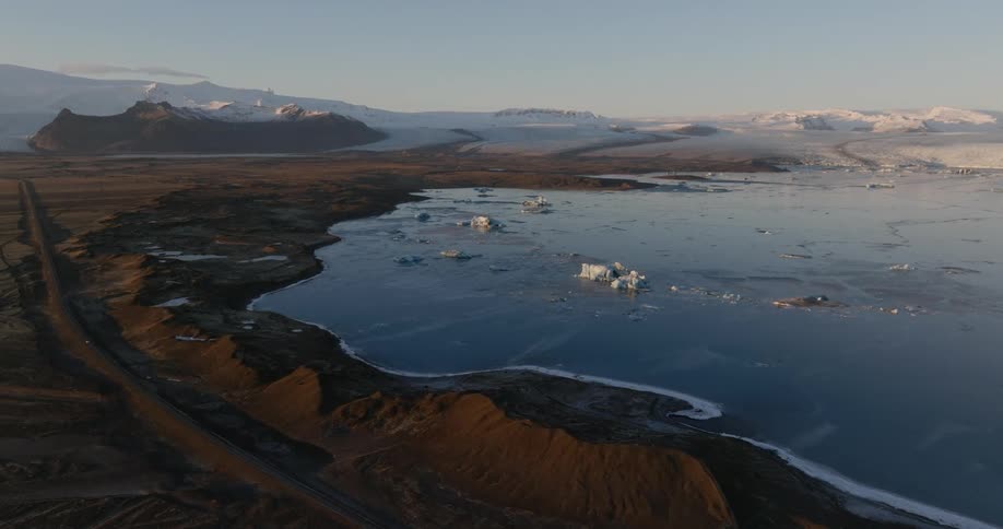 Vehicle moves along road passing shore with nearby icebergs and distant mountains