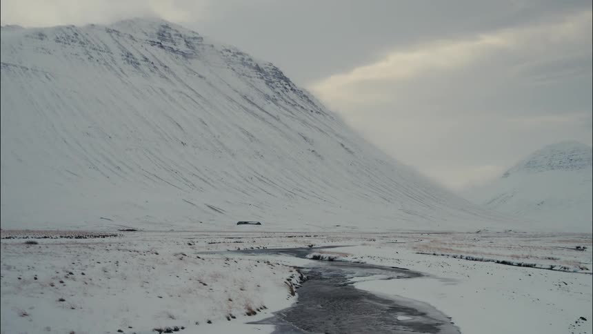 Dark colored river water contrasts with snow covered mountains and fields under an overcast sky