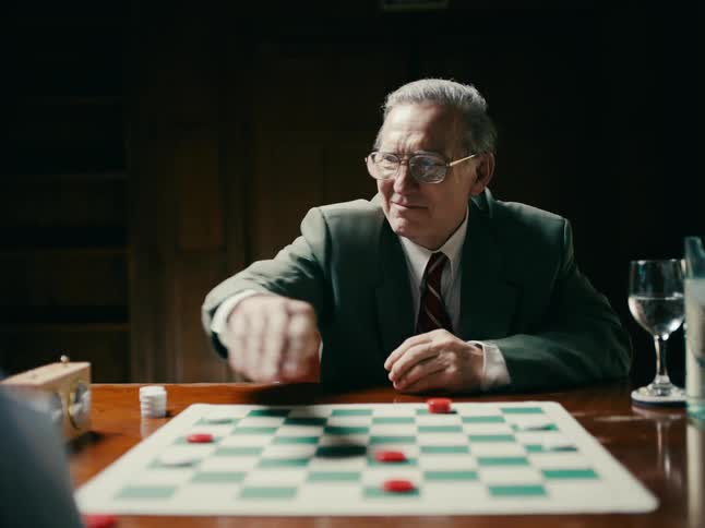 A man is sitting at a table in front of a checker game with his hands clasped watching his opponent move and they each take a turn and shake hands and he takes off his glasses and drinks from a glass