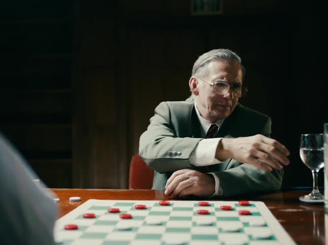 A man is sitting a the table of a checker match with his hands clasped as he waits for his opponent to make a move he takes a drink of water