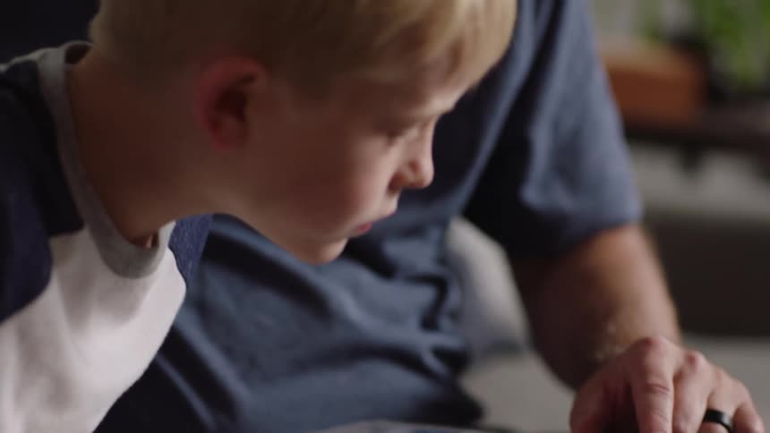 A young boy sits next to his father on a couch and leans over to look at the baseball cards neatly arranged in a binder while his father points at the individual cards with the hand his wedding ring is on