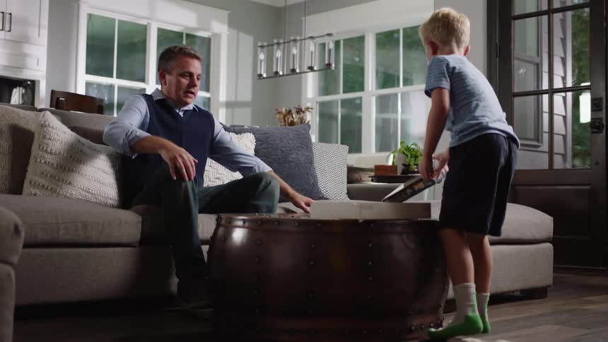 A young boy shakes and opens a puzzle box while his father sits down on the living room couch